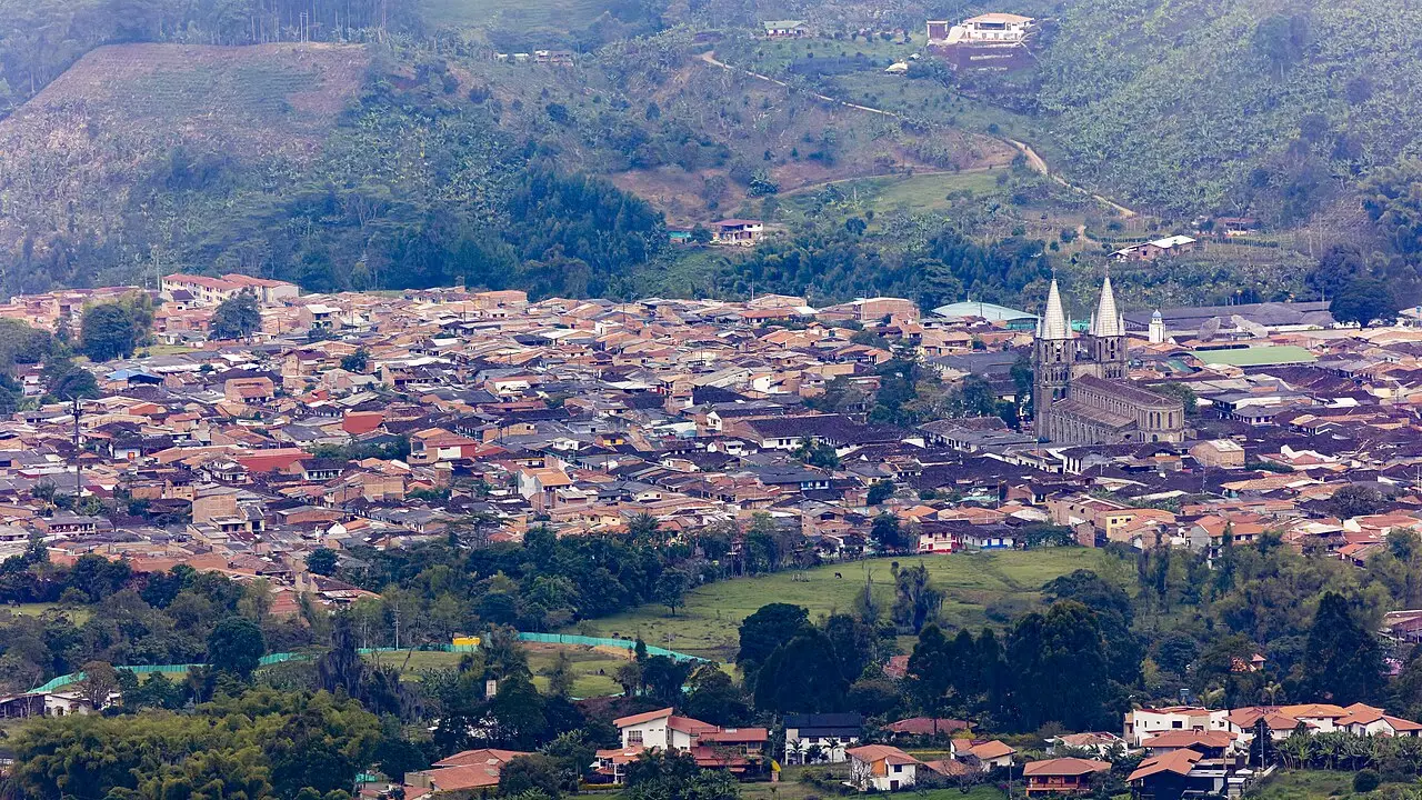 Vista panorâmica de Jardín, com telhados de telha ocupando o vale, as duas torres pontudas da igreja à direita e colinas verdes ao redor.