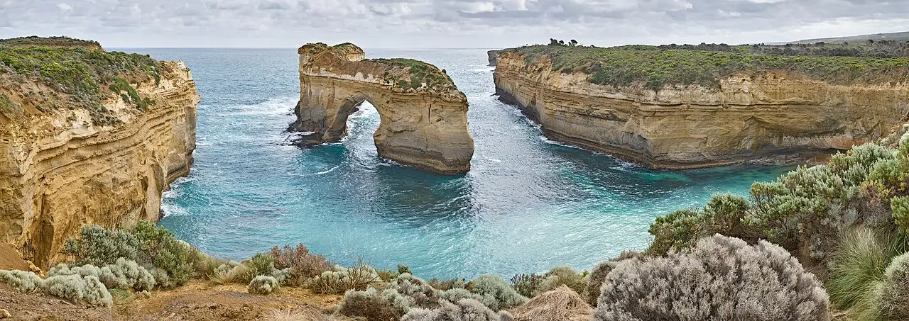 Arco de rocha no mar junto a falésias de arenito amarelado na Great Ocean Road, com água turquesa entre elas e vegetação rasteira em primeiro plano.