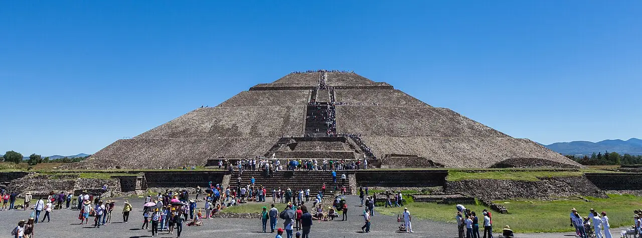 A Pirâmide do Sol em Teotihuacán, com escadaria central, muitos visitantes na base e no topo e céu azul limpo por cima.
