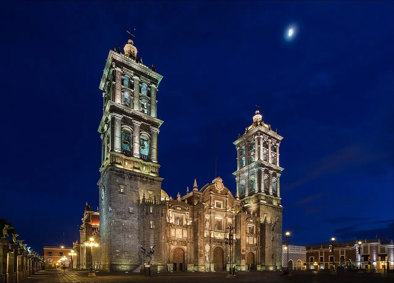 A catedral de Puebla à noite, com as duas torres iluminadas, a fachada de pedra e a praça vazia sob um céu azul escuro com a lua.