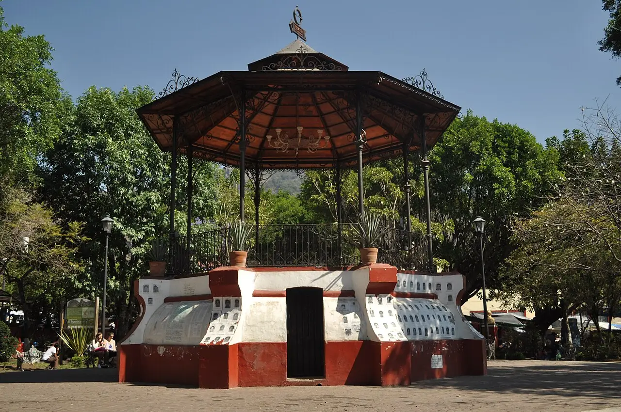 Quiosque de ferro com telhado de cobre octogonal e base branca e vermelha na praça central de Tepoztlán, cercado de árvores altas.