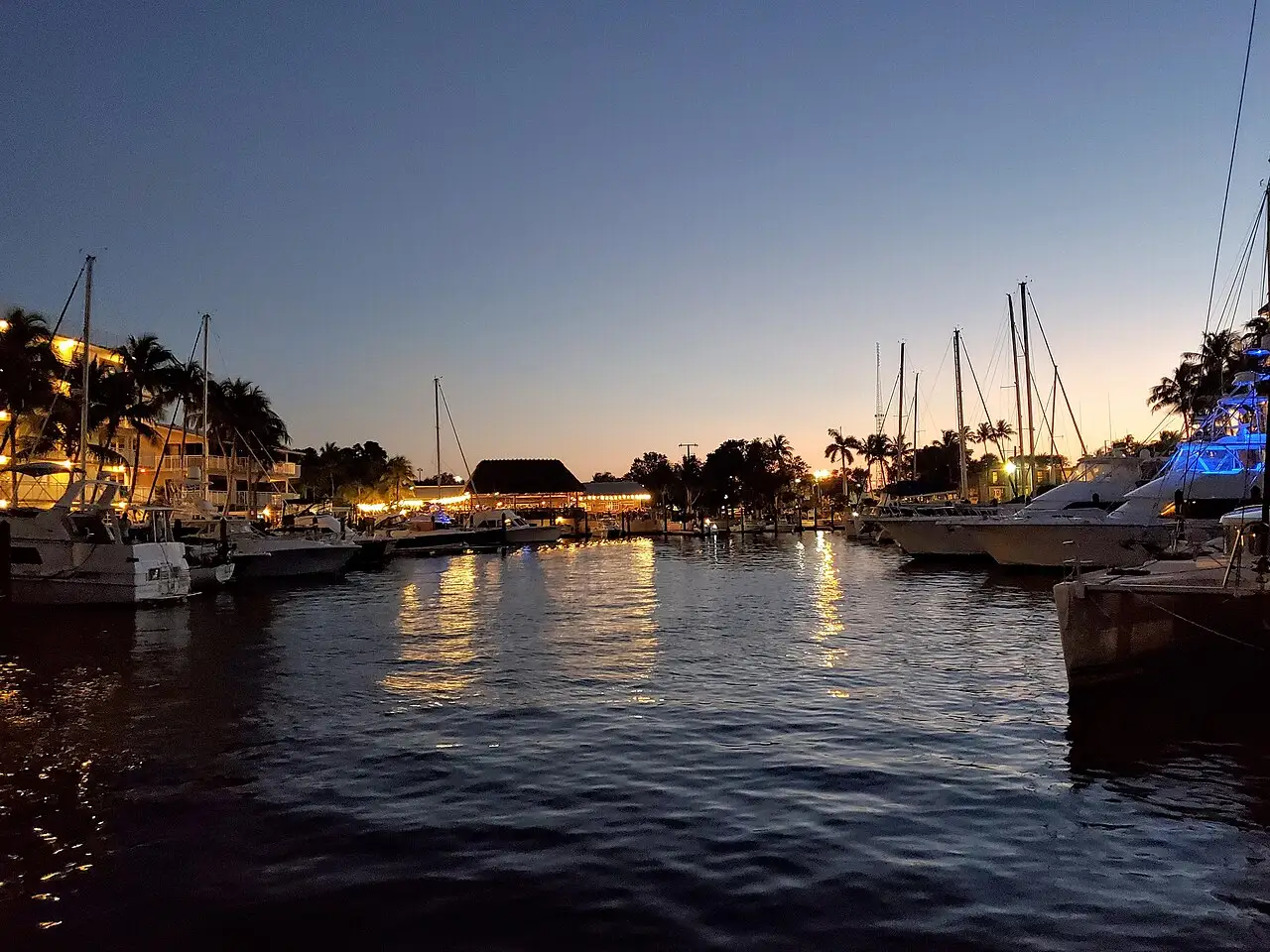 Marina ao anoitecer, com veleiros e as luzes refletidas na água.