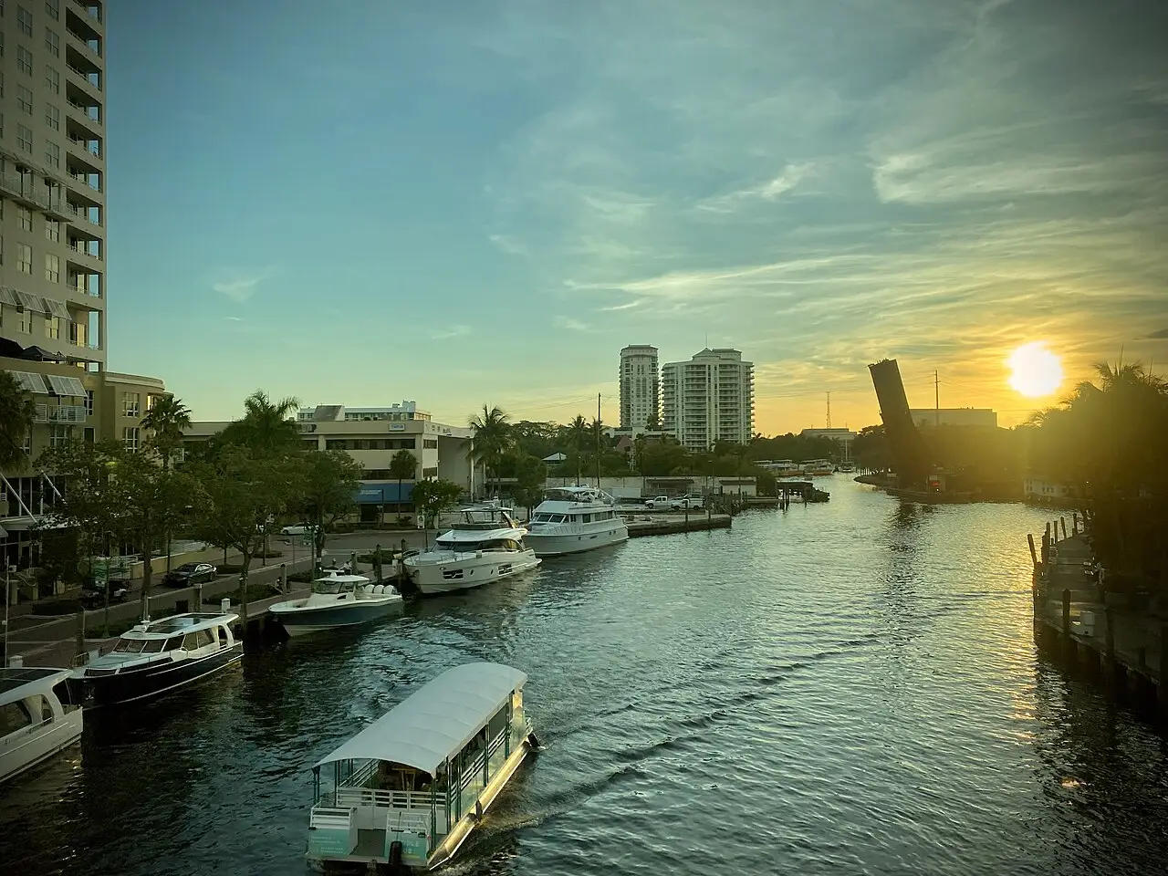 Canal com barcos atracados, o sol baixo e uma ponte levadiça aberta ao fundo.