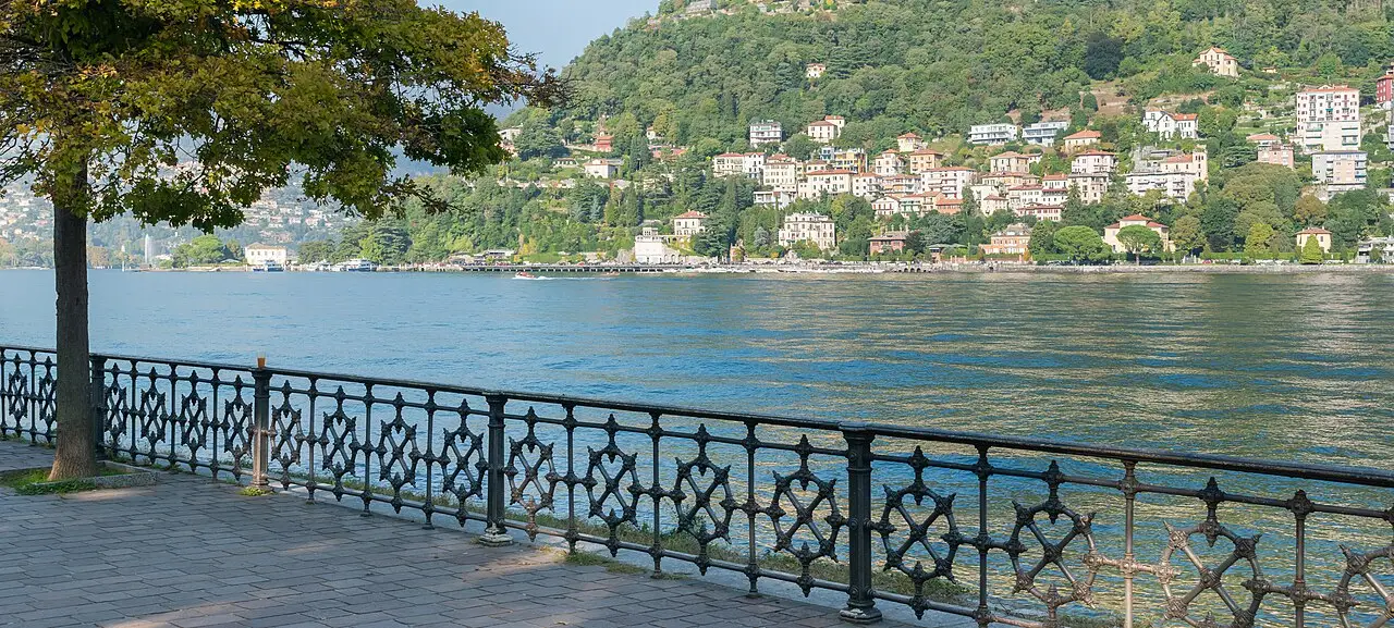 O lago de Como visto da margem de Como, com uma grade de ferro trabalhado em primeiro plano e casas nas encostas verdes da outra margem.
