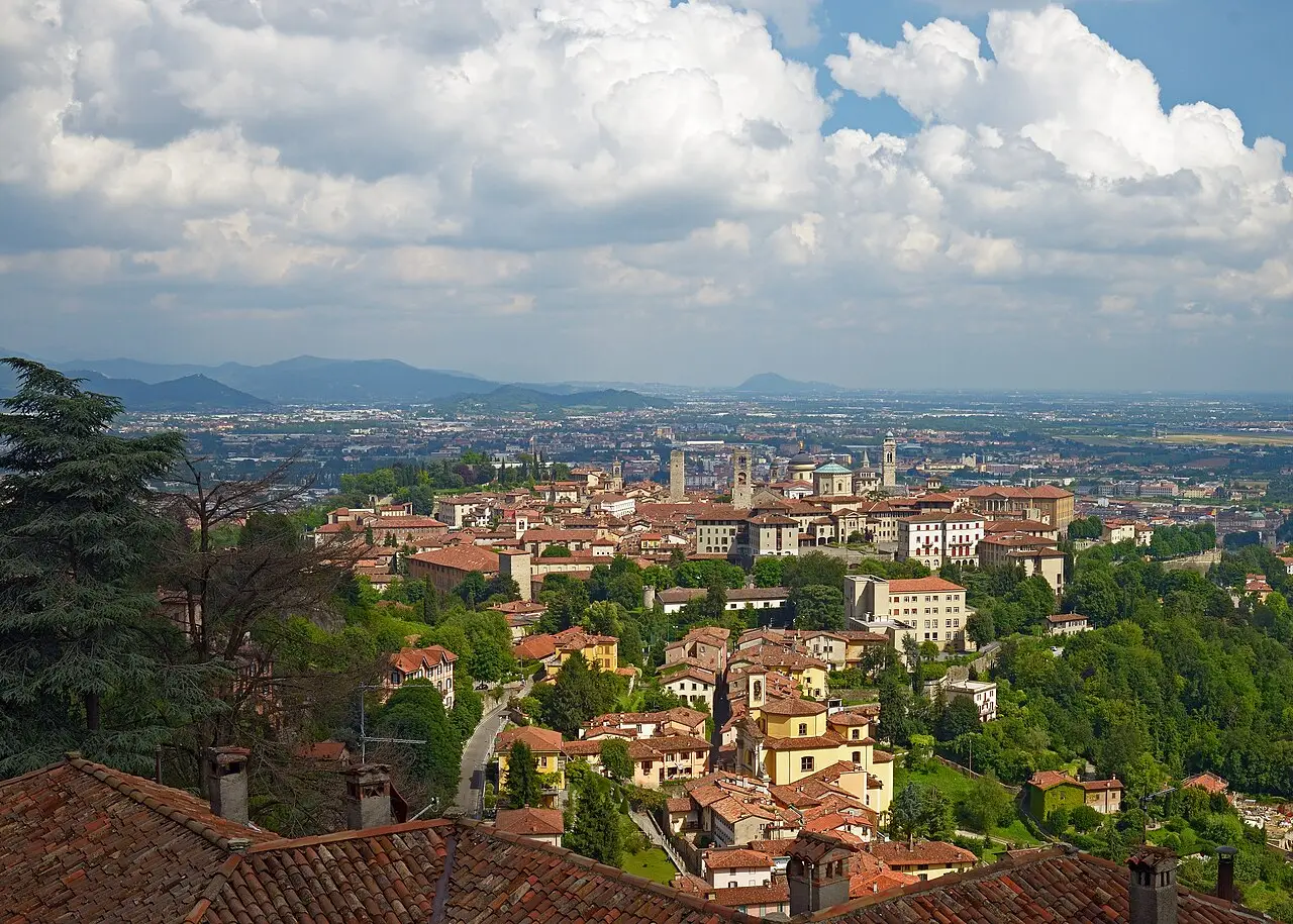 Vista da Cidade Alta de Bergamo entre telhados de telha, com torres e cúpulas ao centro, colinas verdes e a planície ao fundo sob nuvens densas.