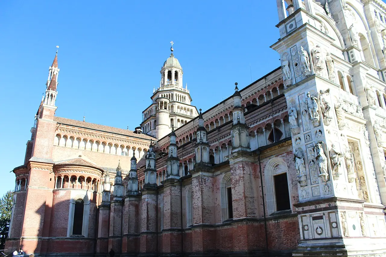 Lateral da Certosa di Pavia, em tijolo e mármore claro, com arcadas, torres e pináculos, a fachada esculpida com estátuas à direita e céu azul.