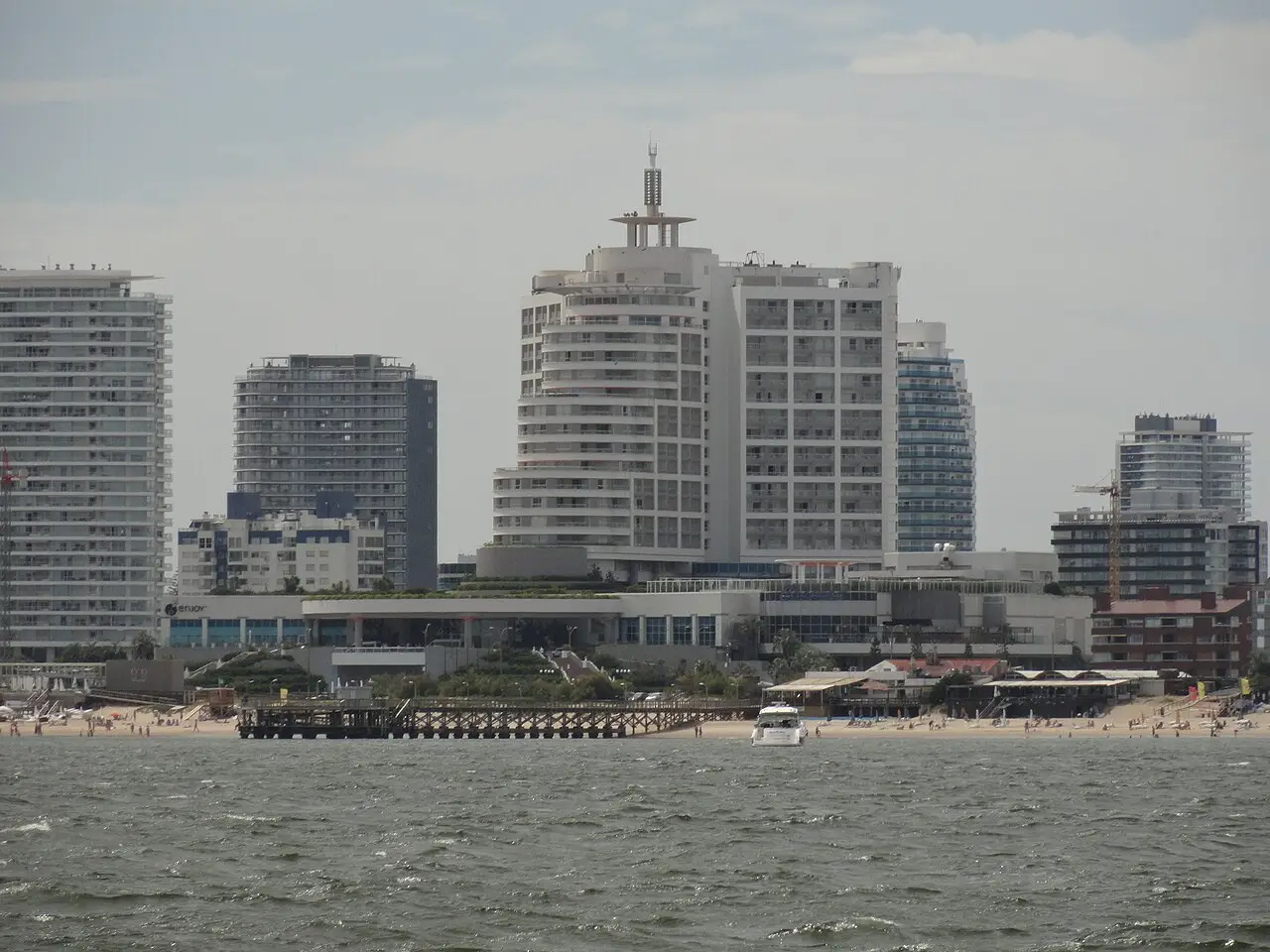 Edifícios altos de apartamentos e hotel na orla de Punta del Este, vistos da água, com uma praia e um píer de madeira na frente.