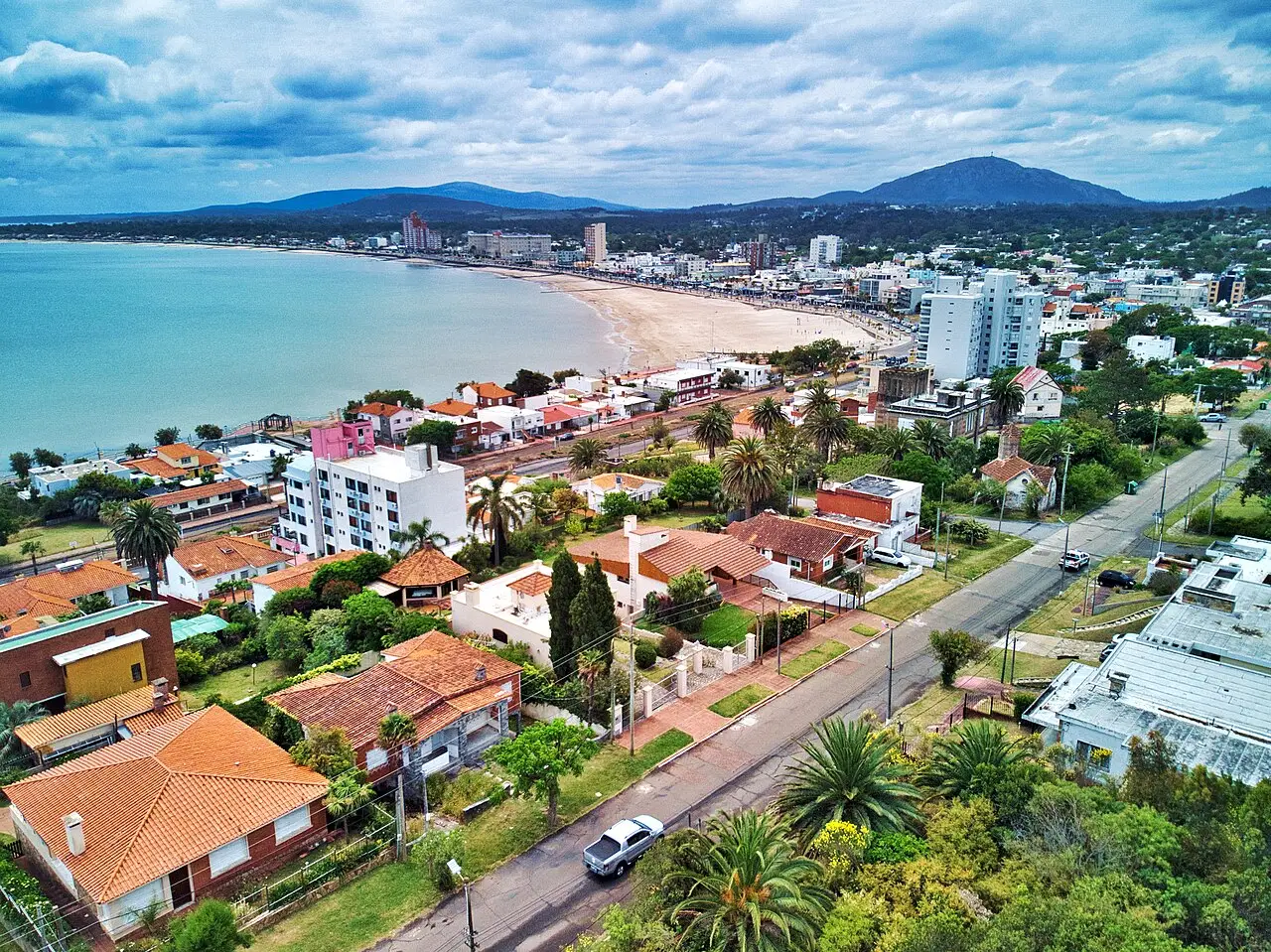 Vista aérea de Piriápolis, com a praia em curva junto a uma baía de água clara, casas de telhado laranja em primeiro plano e serras ao fundo.