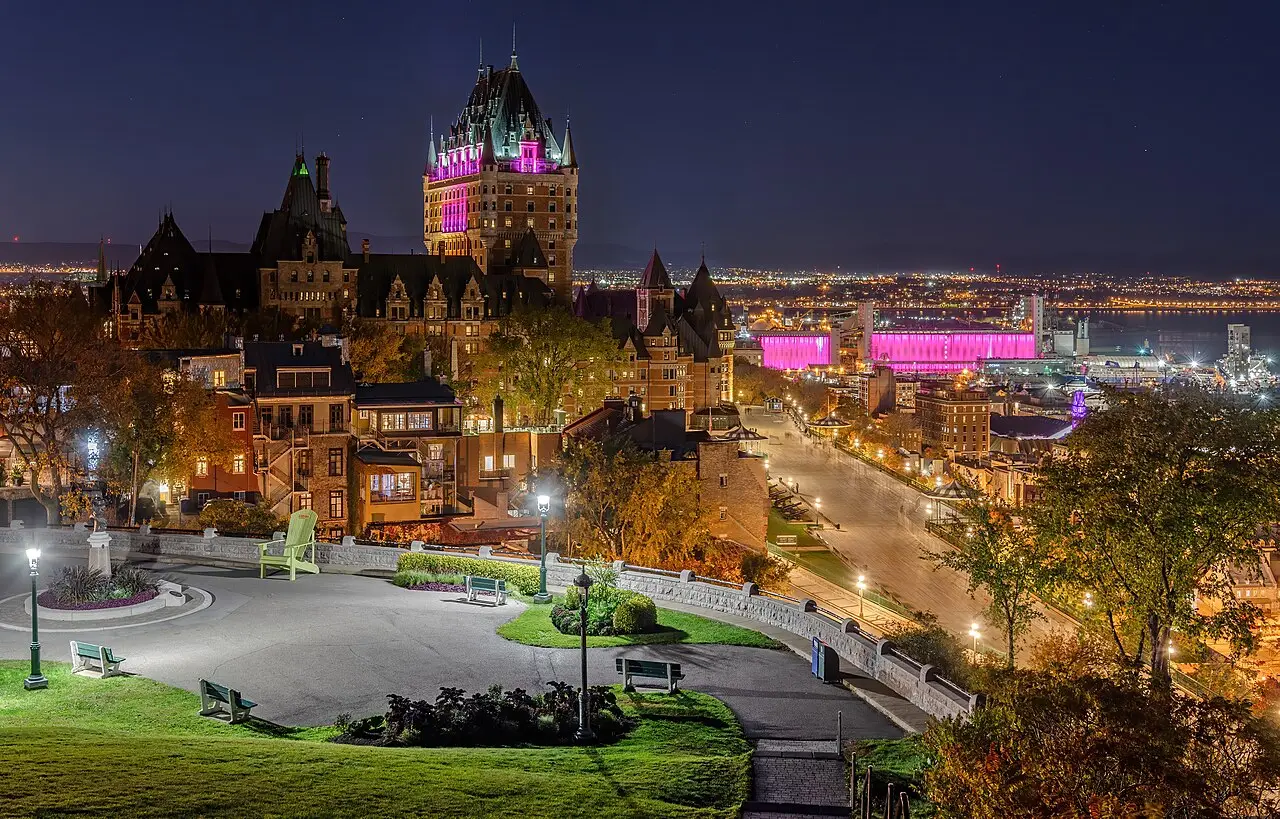 O Château Frontenac à noite, com as torres iluminadas em rosa, o passeio em primeiro plano e as luzes da cidade de Quebec junto ao rio.