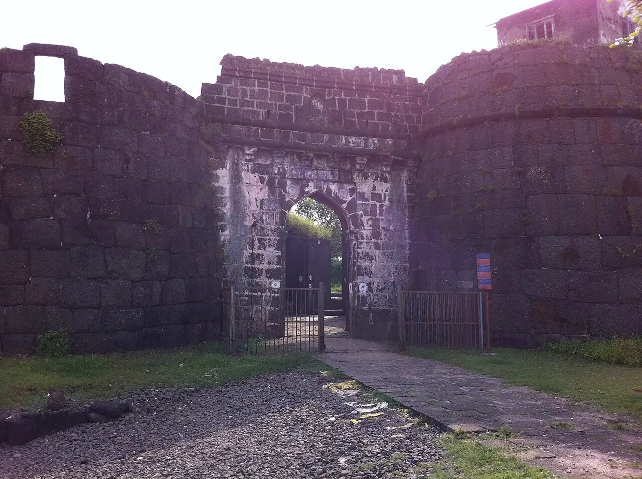 Portão em arco de uma fortificação de pedra escura, entre dois bastiões redondos, com grade de ferro e caminho de pedra à frente, perto de Alibaug.