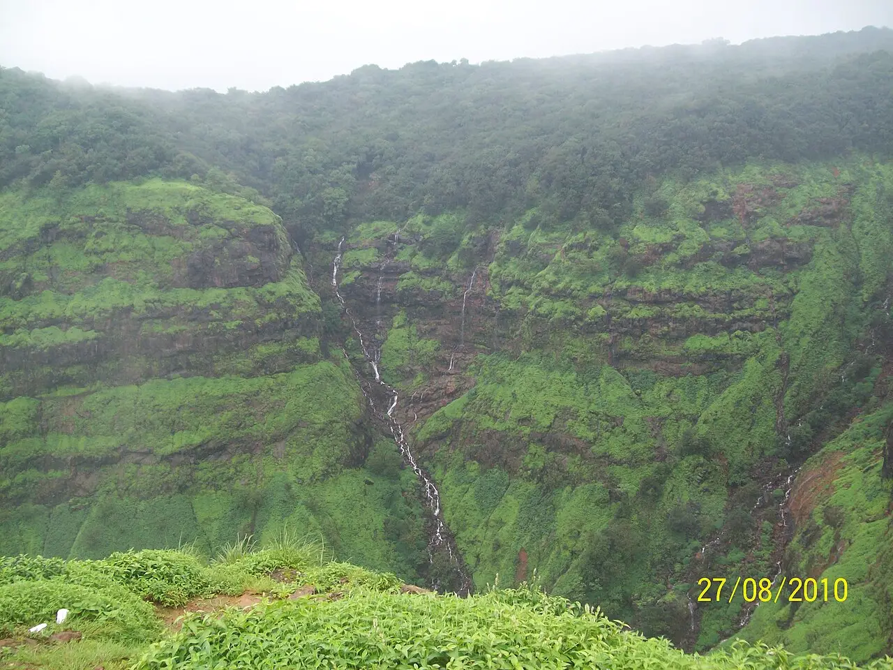Encosta muito verde e enevoada em Matheran, com filetes de cascata descendo pela parede de rocha e mata densa no alto.