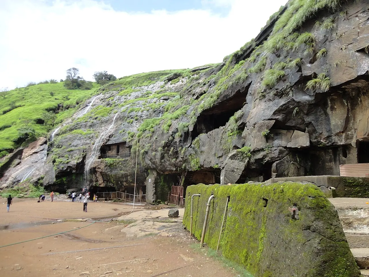 Parede de rocha escura com entradas de cavernas escavadas, cascatas finas e musgo, um terreno de terra batida em frente e visitantes ao longe, perto de Lonavala.
