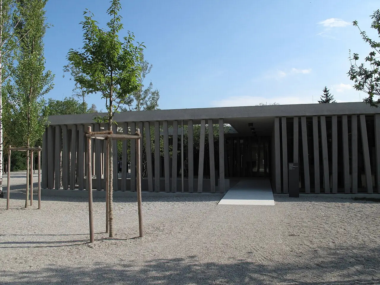 Centro de visitantes do Memorial de Dachau, um edifício baixo de concreto com pilares inclinados, uma passarela clara na entrada e árvores jovens escoradas.