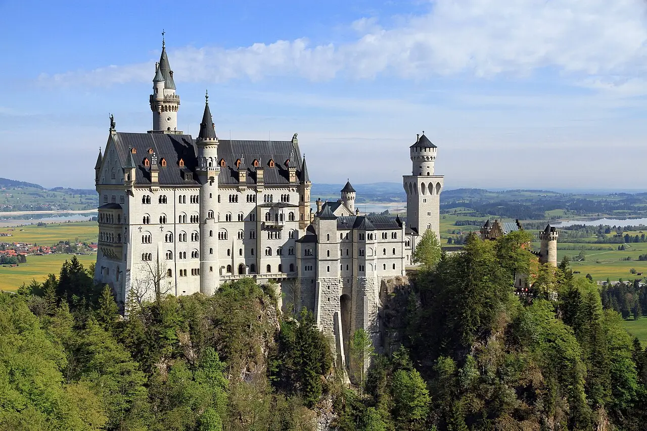 O Castelo de Neuschwanstein sobre uma encosta de floresta, com torres e paredes claras, e campos e lagos na planície ao fundo.
