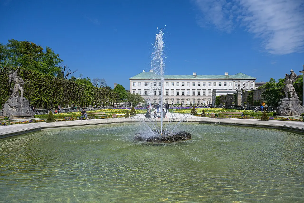 Fonte com jato de água no centro de um tanque, nos jardins Mirabell de Salzburgo, com canteiros floridos, estátuas e o palácio ao fundo.