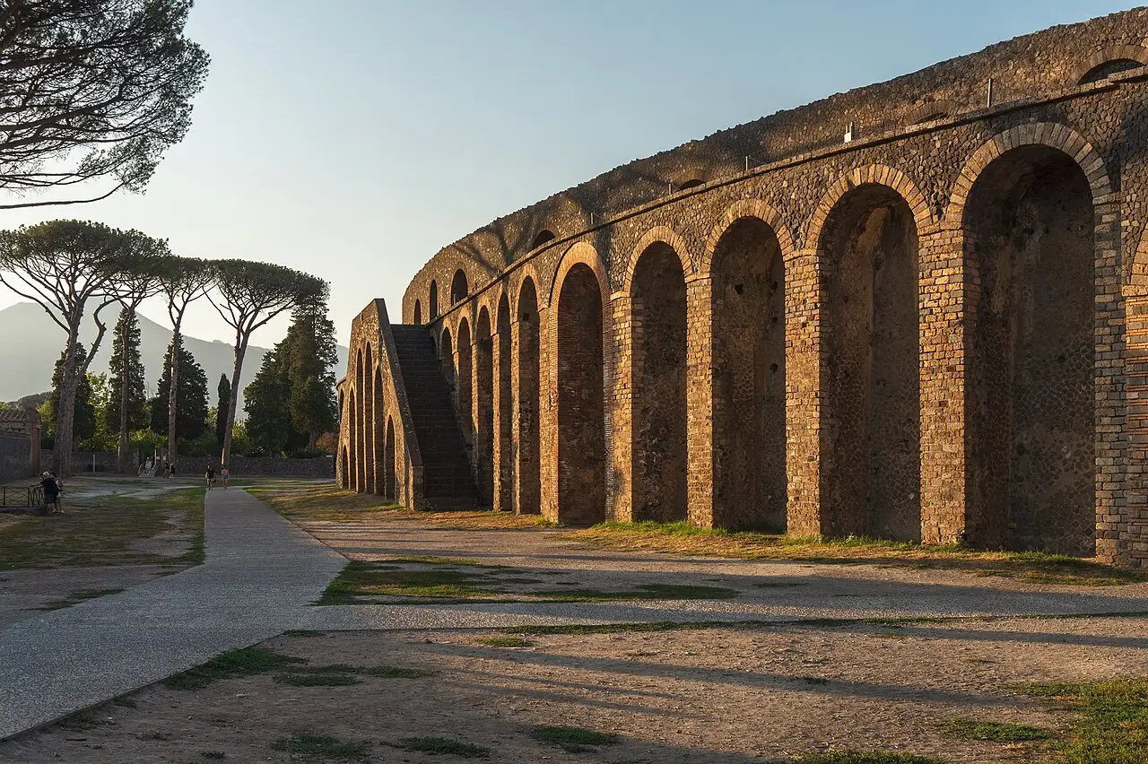 Parede externa em arcos de tijolo e pedra do anfiteatro de Pompeia, com uma escada encostada, pinheiros à esquerda e luz baixa de fim de tarde.