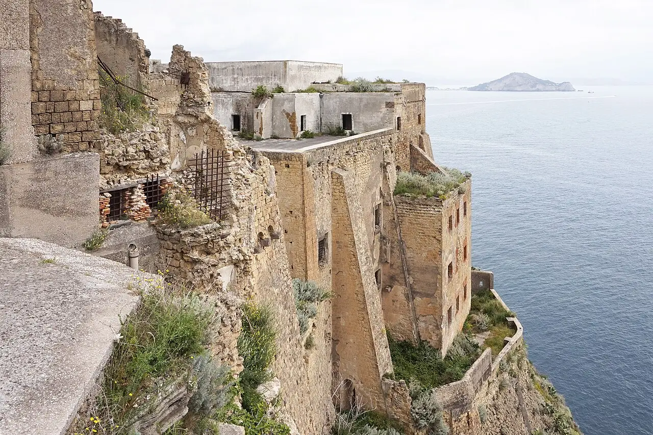 Muros de pedra e tijolo do Palazzo d’Avalos, em Procida, sobre um penhasco junto ao mar, com uma ilha ao longe e vegetação crescendo nas ruínas.