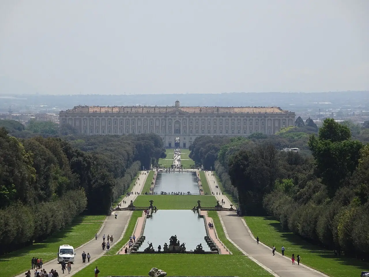 O palácio da Reggia di Caserta visto do fim dos jardins, com canais compridos, alamedas com visitantes e fileiras de árvores dos dois lados.
