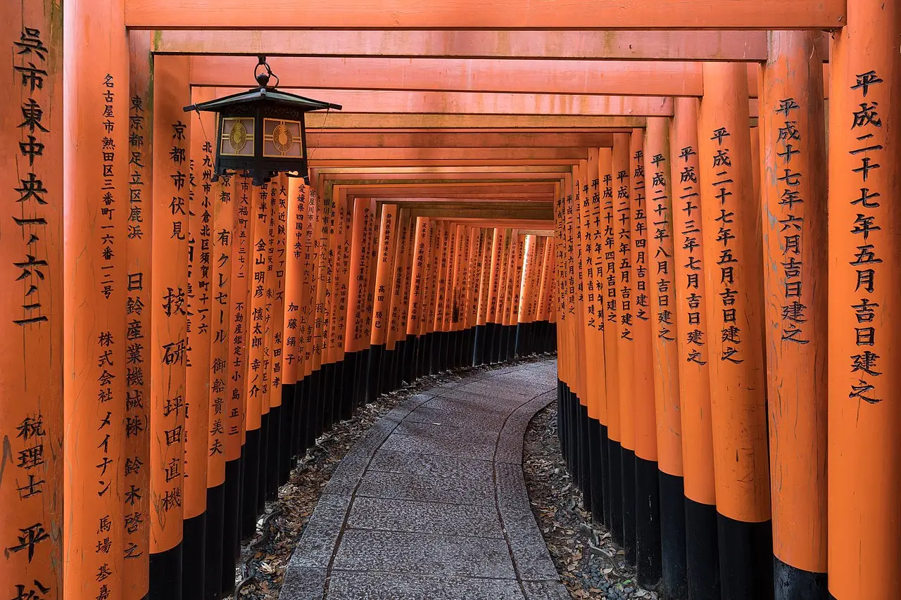 Corredor de portões torii laranja no santuário Fushimi Inari, com inscrições em preto nas colunas, uma lanterna pendurada e um caminho de pedra em curva.