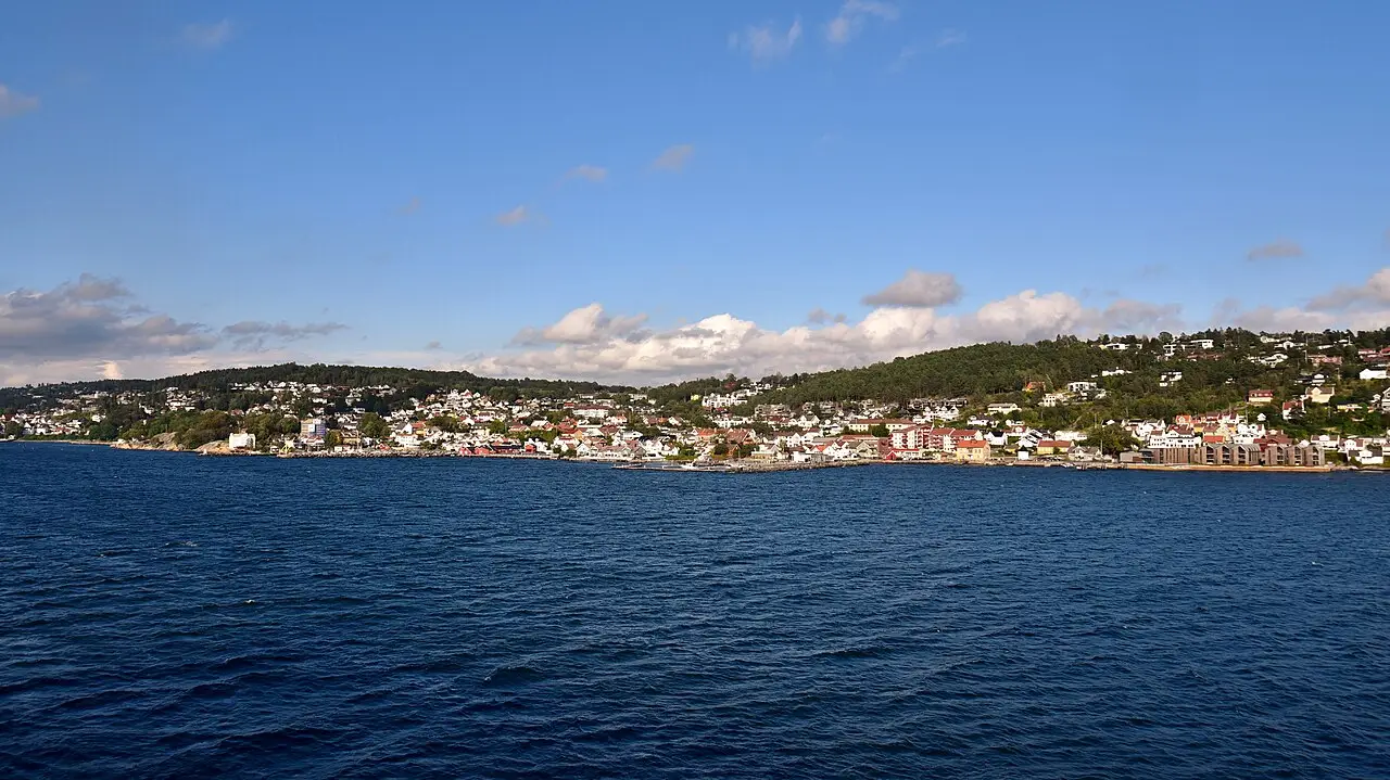 Vue de Drøbak depuis l’eau, avec des maisons blanches et colorées réparties sur la rive basse et boisée, sous un ciel bleu nuageux.