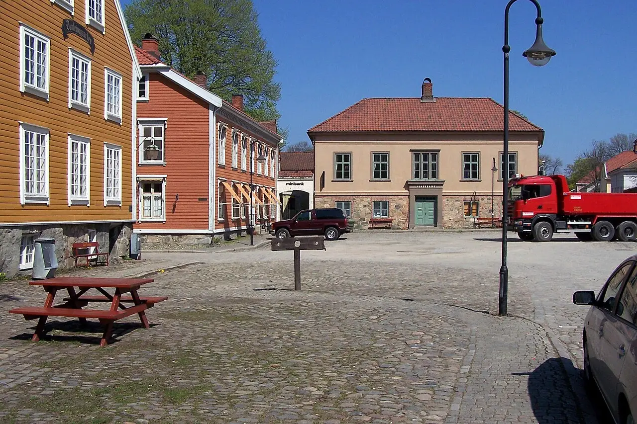Place pavée avec des maisons en bois jaune et orange et un bâtiment beige.