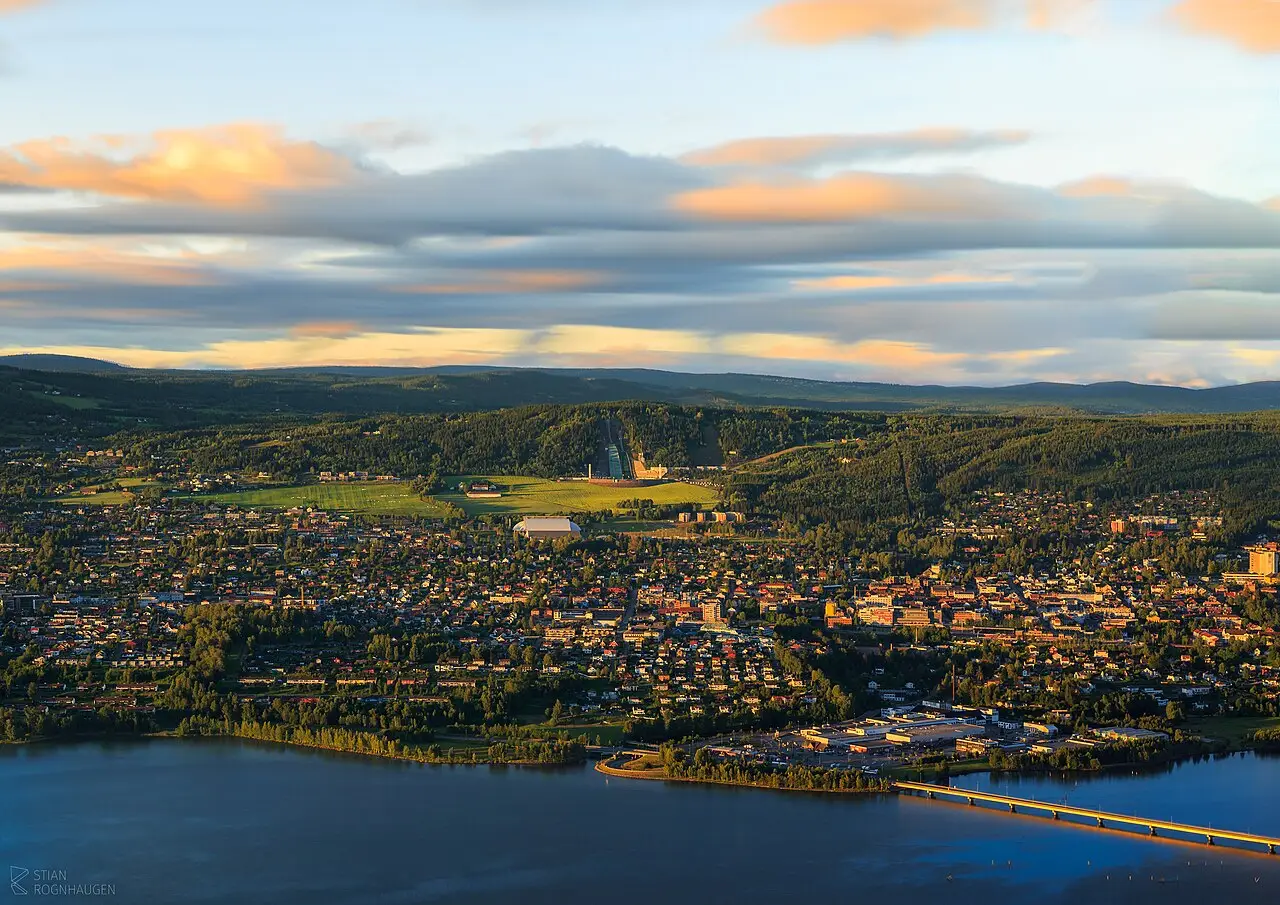 Vue aérienne de Lillehammer au crépuscule, avec la ville entre le lac et les collines boisées, un pont dans le coin et le tremplin de saut à ski au fond.