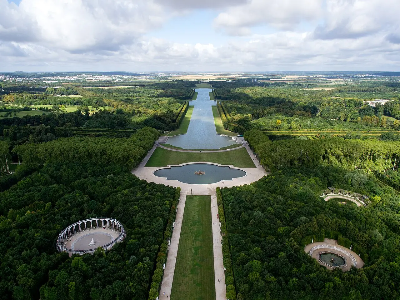 Vista aérea de los jardines de Versalles, con el estanque de Apolo en primer plano, el Gran Canal al fondo y bosquetes de árboles a ambos lados.
