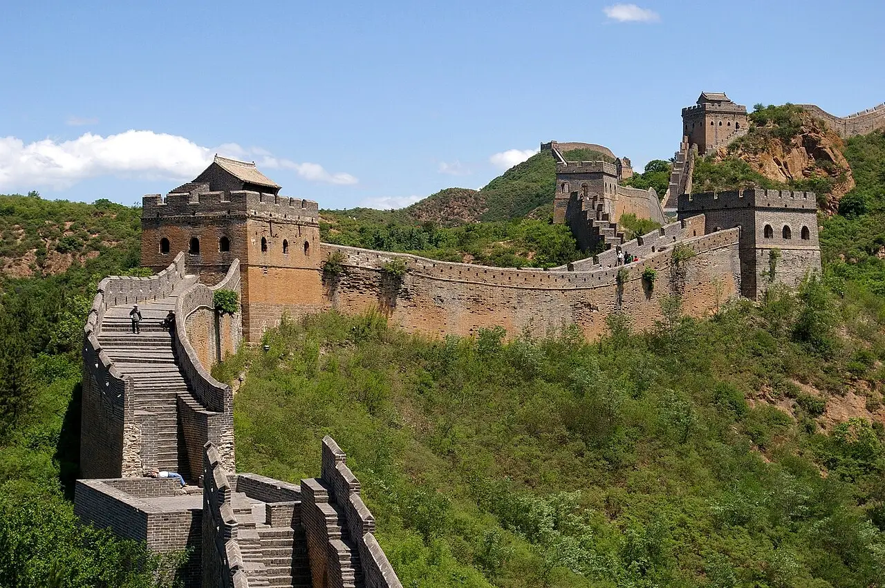 Trecho da Grande Muralha com torres de vigia de tijolo, escadas e ameias, seguindo pela crista de colinas verdes sob céu azul, perto de Jinshanling.