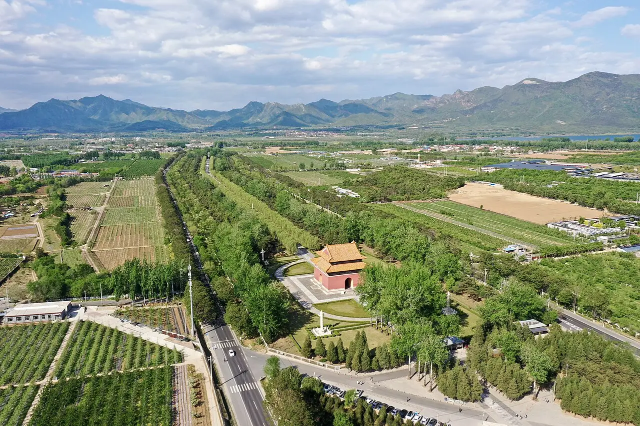 Vista aérea de uma alameda arborizada que passa por um pavilhão de paredes vermelhas e telhado amarelo, com campos cultivados e montanhas ao fundo.