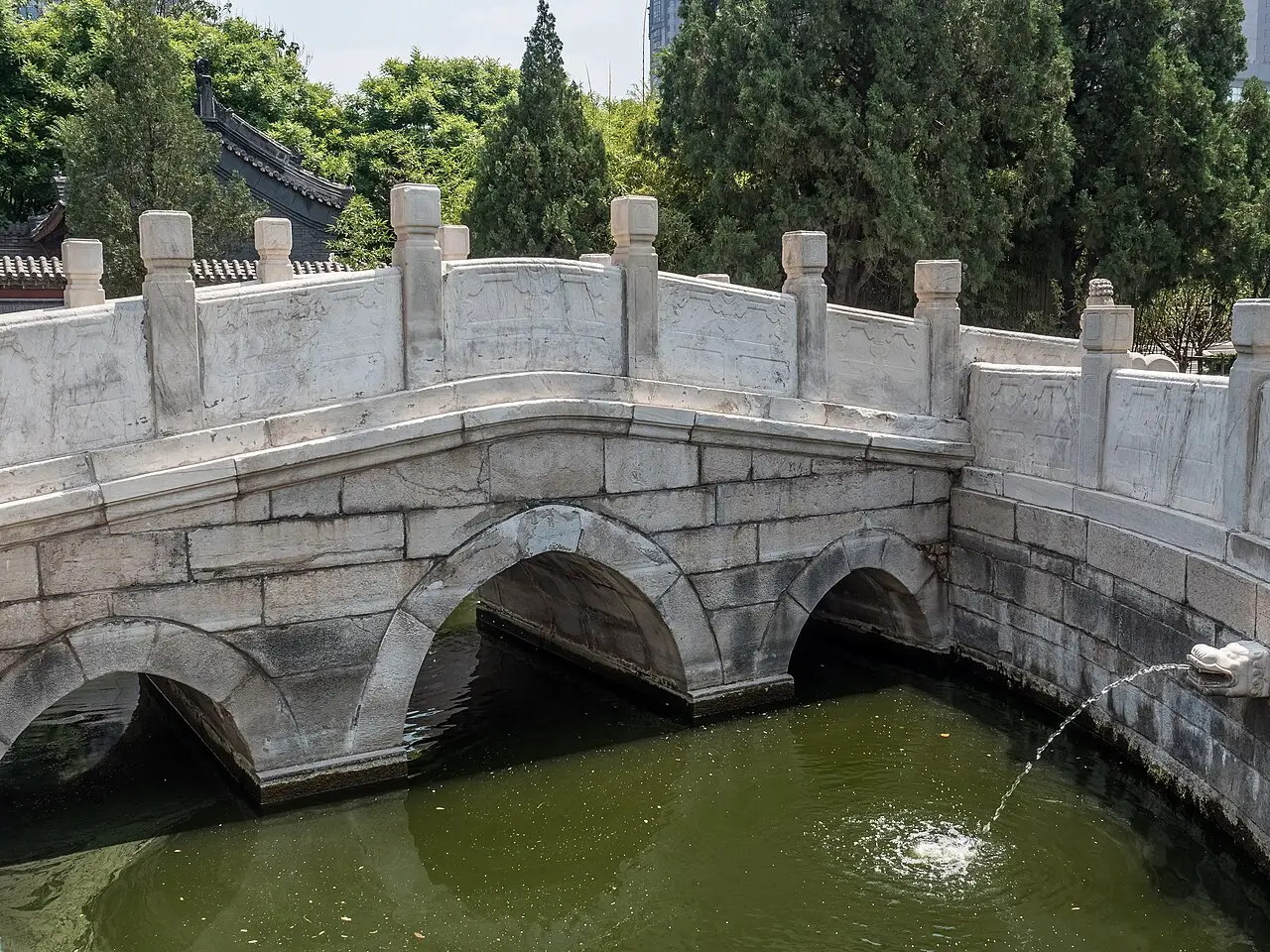 Ponte de pedra branca com três arcos sobre um canal de água esverdeada, com balaustrada esculpida, uma cabeça de dragão de onde jorra água e árvores ao fundo, em Tianjin.