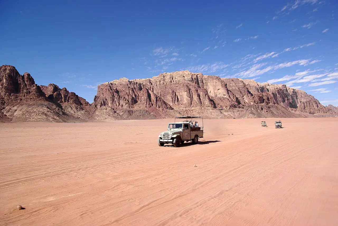 Caminhonete aberta atravessa a planície de areia rosada do Wadi Rum, com outros veículos ao longe e paredões de arenito vermelho sob céu azul.