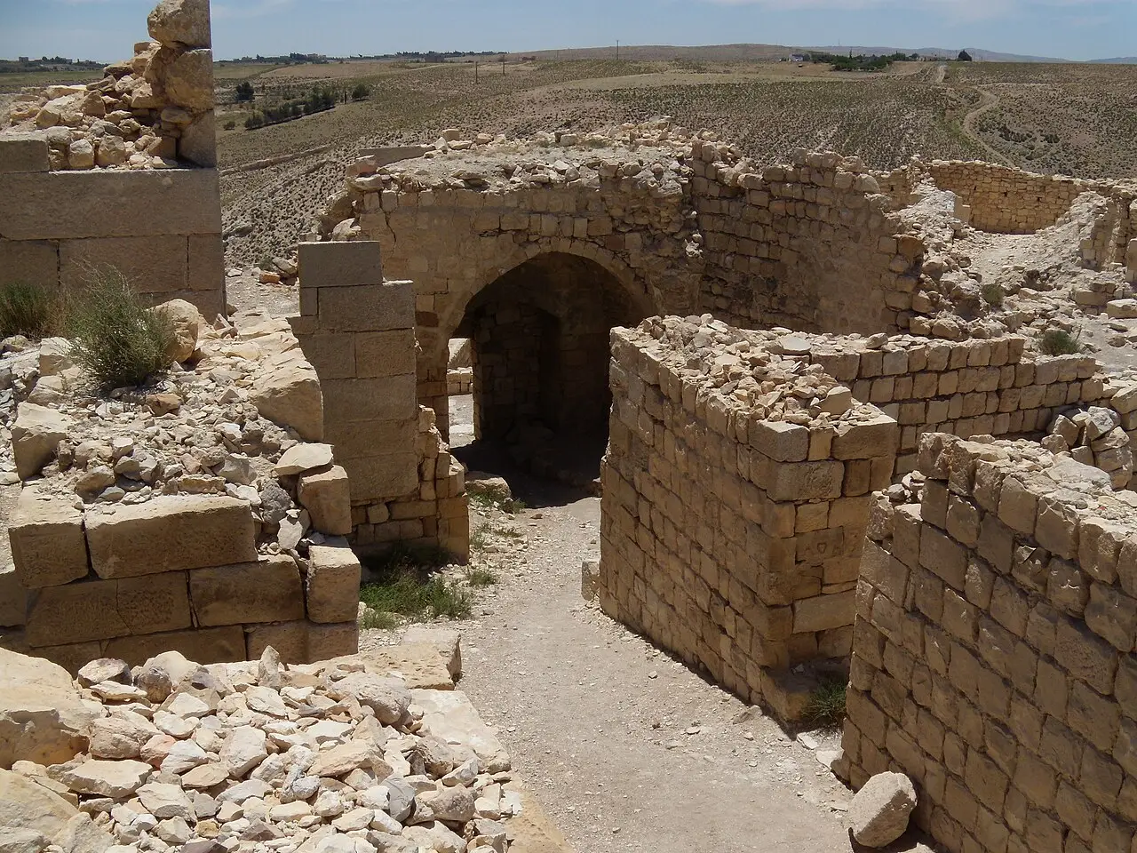 Ruínas de pedra clara do castelo de Shobak, com uma passagem em arco entre muros baixos e campos secos e planos ao fundo.