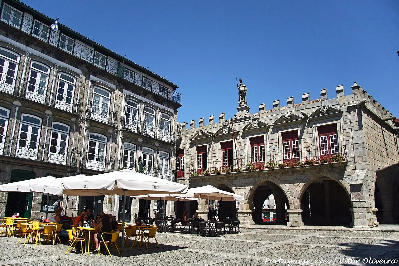 Praça com casas de granito, o antigo edifício com arcos e ameias à direita, uma estátua no topo e mesas de café com guarda-sóis à esquerda, em Guimarães.