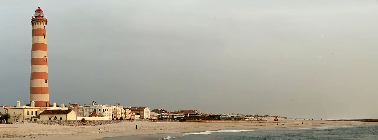 Farol alto de listras laranja e brancas junto a uma fileira de casas, com a praia de areia e o mar em primeiro plano sob céu cinzento, em Aveiro.