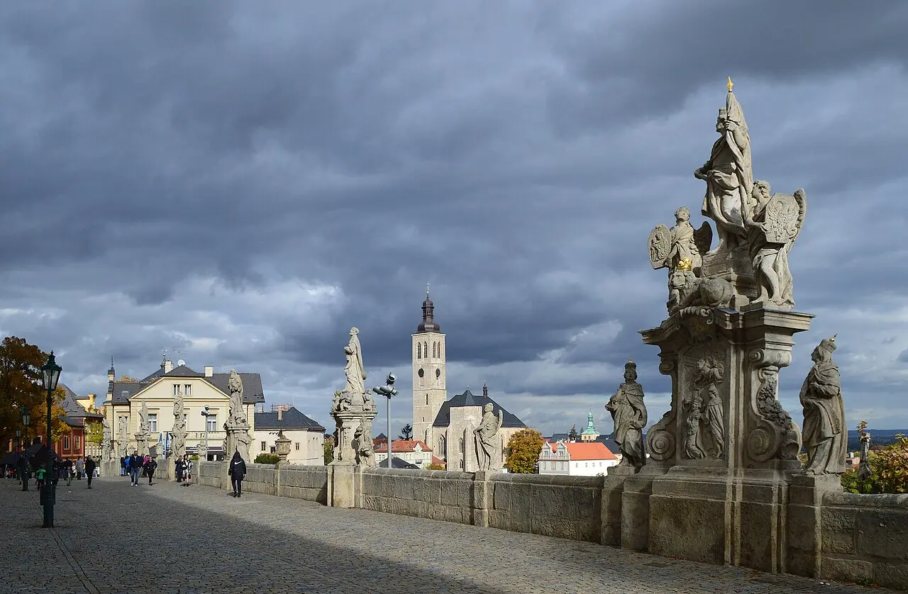 Balaustrada com grupos de estátuas barrocas de pedra em primeiro plano, a torre da igreja de São Tiago ao centro e casas de telhado laranja, em Kutná Hora.