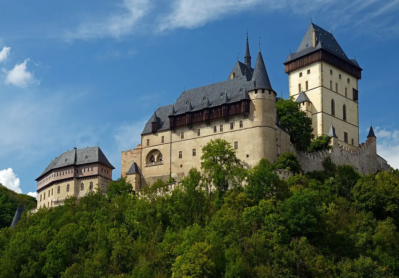 O castelo de Karlštejn no alto de uma colina coberta de árvores, com torres de telhado escuro, muralha com ameias e céu azul.