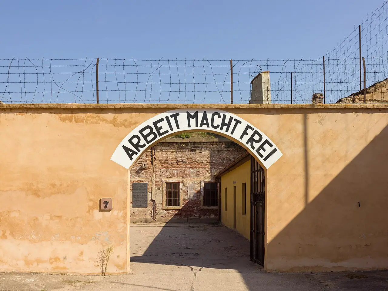 Portão em arco de um muro ocre com a inscrição Arbeit macht frei, arame farpado no alto e um pátio de tijolo ao fundo, em Terezín.