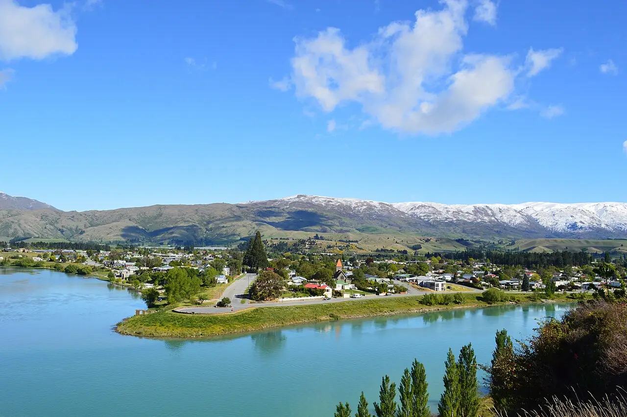 A cidade de Cromwell junto ao rio de água turquesa, com casas entre árvores e montanhas com neve nos picos ao fundo, sob céu azul.