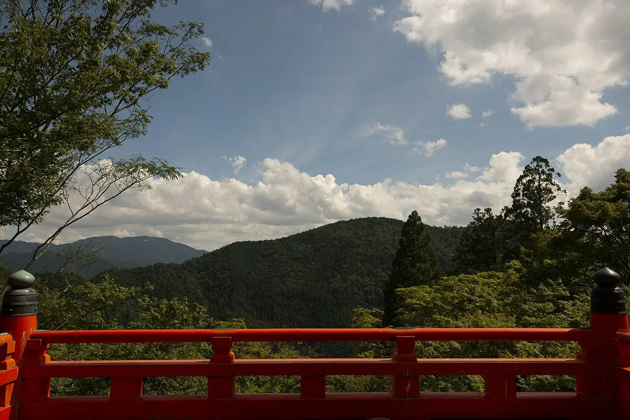 Balaustrada vermelha de uma varanda do Kurama-dera, com vista para colinas cobertas de floresta e um céu com nuvens grandes.