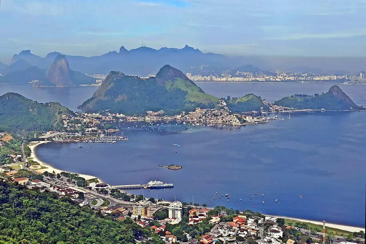 Vista do alto da baía de Niterói, com praia e barcas, uma península de casas ao fundo, morros verdes e, do outro lado da água, a silhueta do Rio de Janeiro.