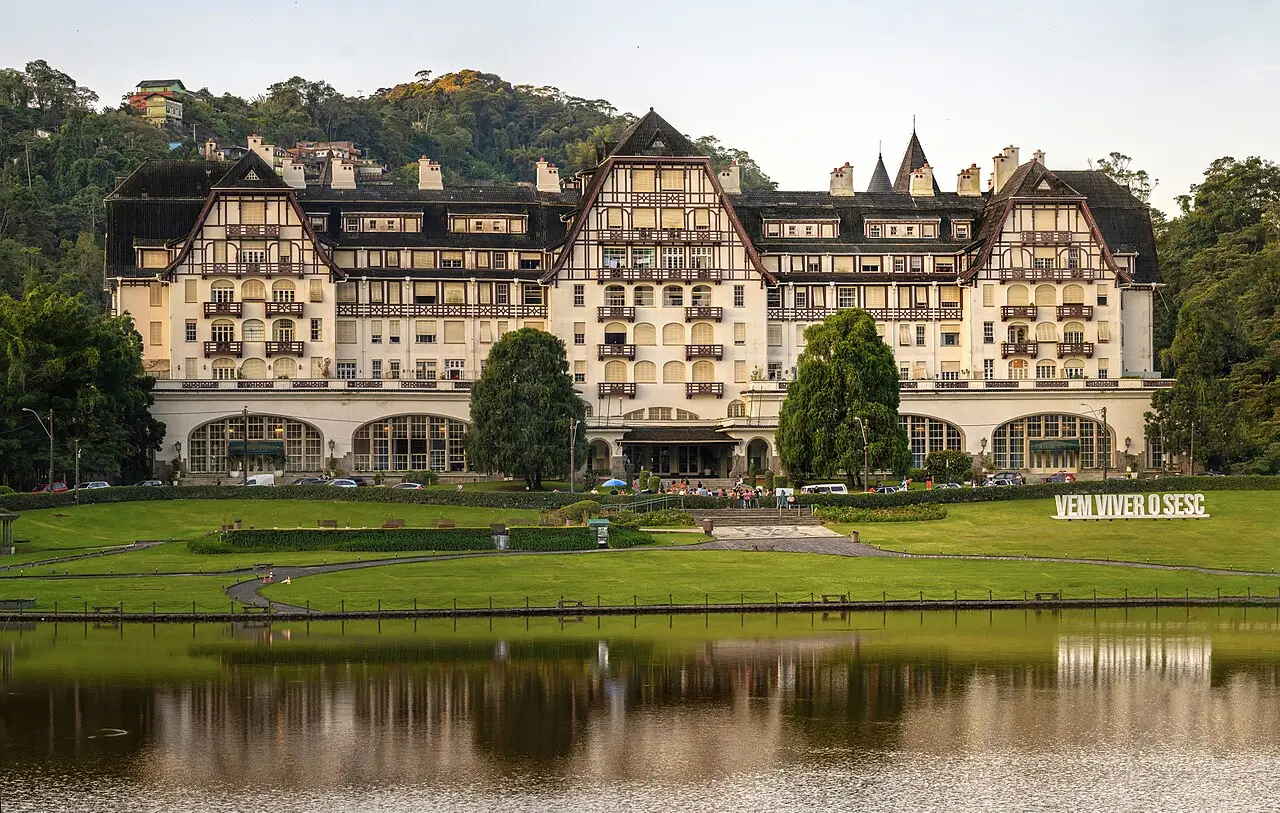 O Palácio Quitandinha, grande edifício de estilo enxaimel com telhados escuros, atrás de um gramado e de um lago que reflete a fachada, em Petrópolis.
