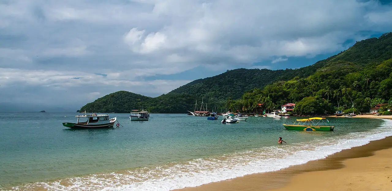 Praia de areia dourada com ondas suaves e barcos ancorados numa enseada de água esverdeada, com morros cobertos de mata e casas na beira, em Ilha Grande.