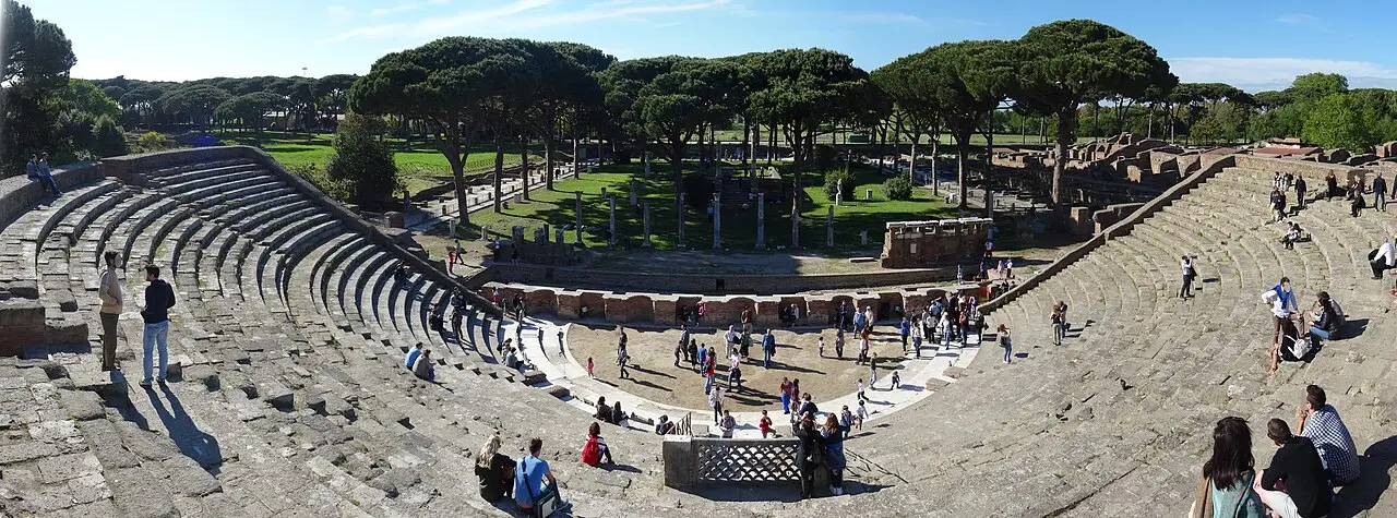 Panorama do teatro de Ostia Antica, com as arquibancadas de pedra dos dois lados, o palco no centro com visitantes e pinheiros ao fundo.