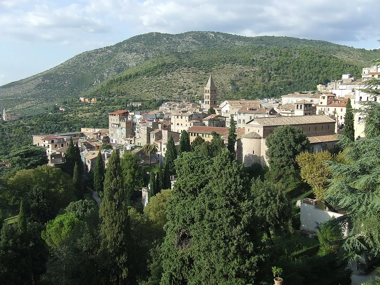 Vista da cidade antiga de Tivoli a partir dos jardins da Villa d’Este, com cipreste em primeiro plano, torre de igreja, casas de telhado de telha e uma serra ao fundo.