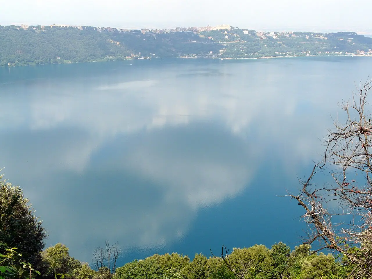 Vista ampla do lago Albano, cercado por encostas verdes, com Castel Gandolfo no alto da margem e nuvens refletidas na água.