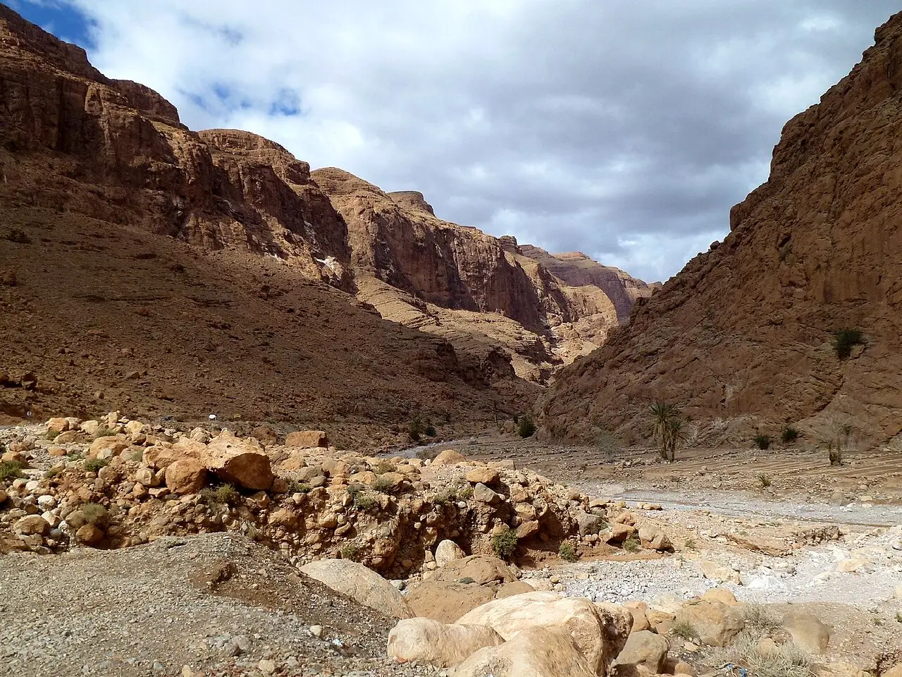 Un desfiladero de altas paredes de roca rojiza, con un lecho seco de río cubierto de piedras y algunas palmeras al fondo, bajo las nubes.