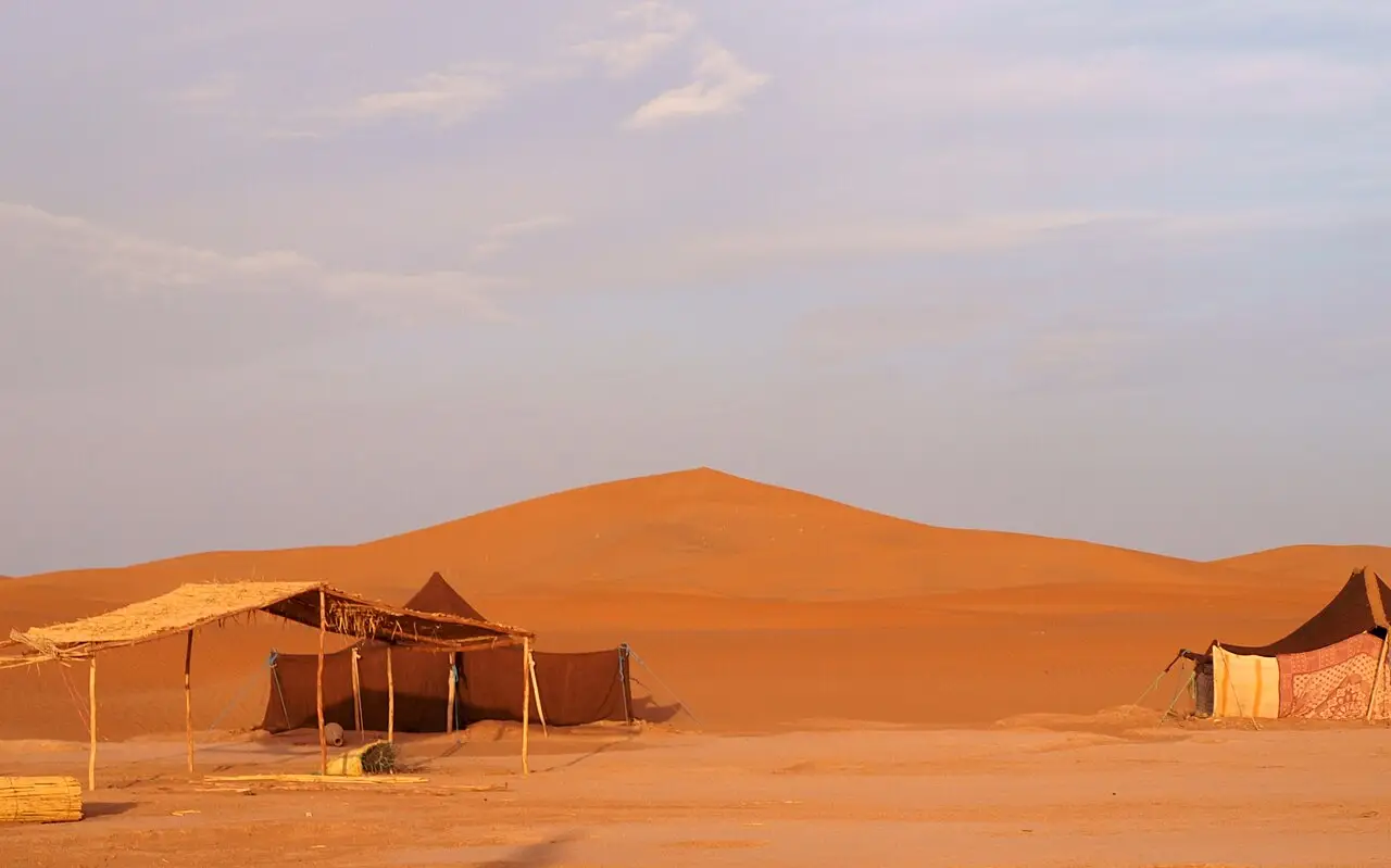 Tendas simples de tecido escuro e cobertura de palha ao pé de uma grande duna de areia alaranjada, na luz do amanhecer, em Erg Chigaga.