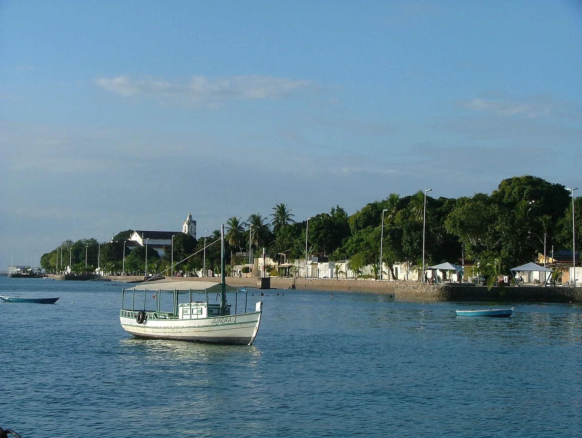 Um barco pequeno na água, com a margem e as casas atrás.