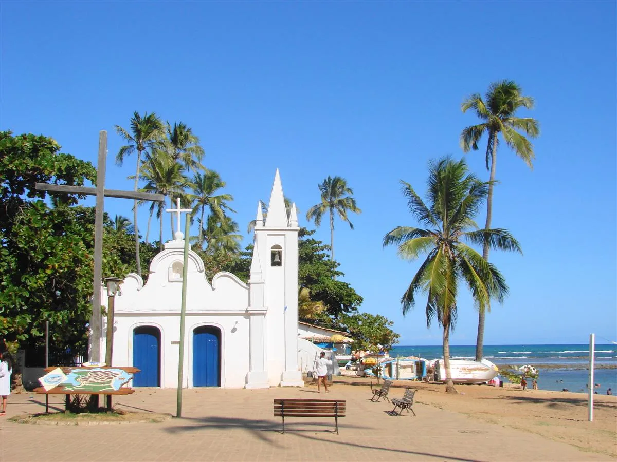Igreja branca de portas azuis, com palmeiras e o mar ao lado.