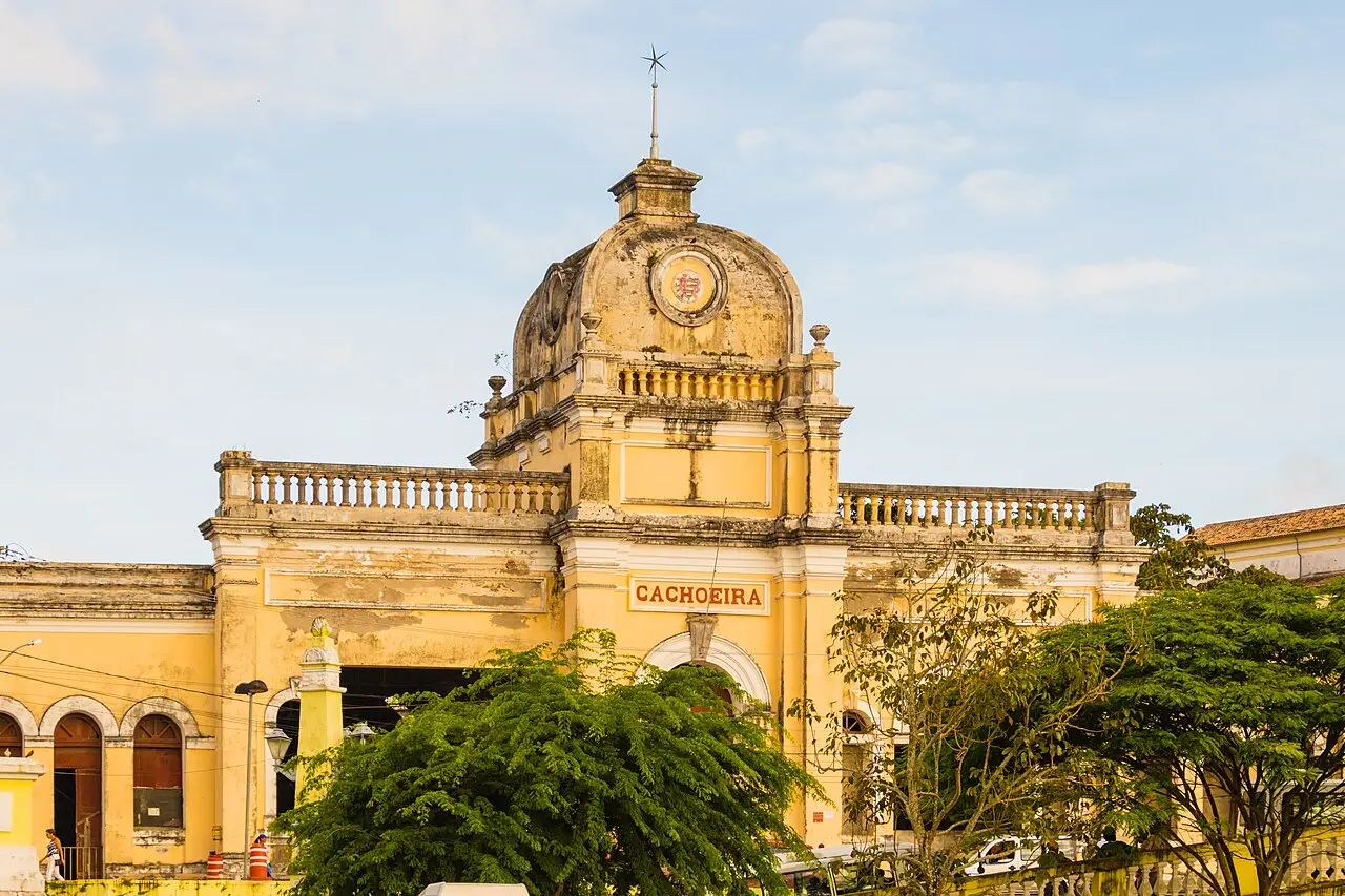 Fachada amarela da antiga estação ferroviária de Cachoeira, com a palavra Cachoeira sobre a entrada, balaustradas e árvores em primeiro plano.